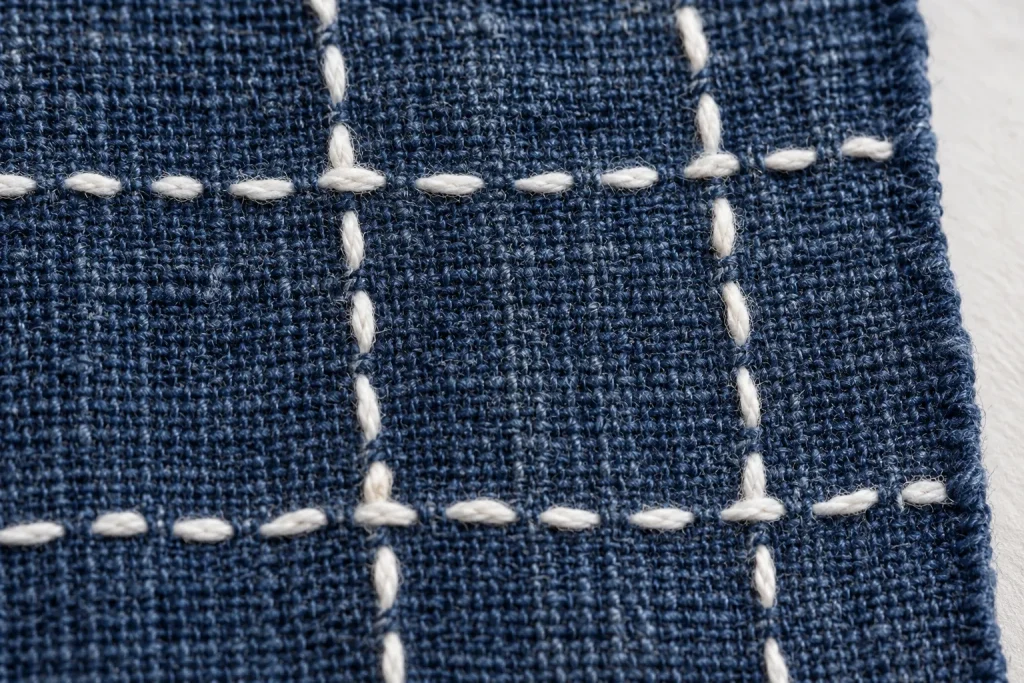 A close view of white counted stitches crossing indigo bast-fiber cloth, with the weave clearly visible
